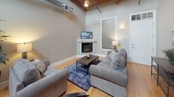 This two-story space blends industrial character with comfortable living, showcasing an open layout anchored by soaring ceilings with exposed wood beams, visible ductwork, and light hardwood floors throughout. The living area is a welcoming focal point, centered around a corner fireplace and furnished with ample seating. A dramatic black spiral staircase connects the main level to the upper loft, serving as both a functional element and a striking architectural feature.

The kitchen is well-appointed with warm wood cabinetry, granite countertops, stainless steel appliances, and a breakfast bar that opens to the dining and living areas, making it ideal for both everyday use and entertaining. Two bedrooms on the main level offer a calm retreat, each featuring hardwood floors and neutral tones, with dark wood furniture providing a classic contrast. The full bathroom includes tile flooring, a granite-topped vanity, and a tub-shower combination.

The upper loft is a standout feature, boasting an arched wood-plank ceiling, exposed timber framing, and multi-pane windows on all sides that flood the space with natural light, creating a bright and versatile bonus area.
