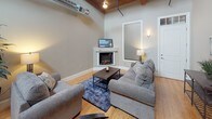 This two-story space blends industrial character with comfortable living, showcasing an open layout anchored by soaring ceilings with exposed wood beams, visible ductwork, and light hardwood floors throughout. The living area is a welcoming focal point, centered around a corner fireplace and furnished with ample seating. A dramatic black spiral staircase connects the main level to the upper loft, serving as both a functional element and a striking architectural feature.

The kitchen is well-appointed with warm wood cabinetry, granite countertops, stainless steel appliances, and a breakfast bar that opens to the dining and living areas, making it ideal for both everyday use and entertaining. Two bedrooms on the main level offer a calm retreat, each featuring hardwood floors and neutral tones, with dark wood furniture providing a classic contrast. The full bathroom includes tile flooring, a granite-topped vanity, and a tub-shower combination.

The upper loft is a standout feature, boasting an arched wood-plank ceiling, exposed timber framing, and multi-pane windows on all sides that flood the space with natural light, creating a bright and versatile bonus area.