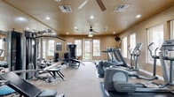 The fitness center at 100 Penzance Ave in Chico, California is a dedicated, single-room community gym designed to support a well-rounded exercise routine. The space features warm, neutral-toned walls paired with light carpeting, creating an inviting and energizing atmosphere. Ceiling fans with integrated lighting and recessed can lights ensure the room is well-lit and comfortable throughout the day.

Natural light fills the space through multiple windows positioned along the walls, as well as a glass-paneled entry door that opens to the surrounding grounds, offering pleasant outdoor views. The room is efficiently organized into distinct cardio and strength training zones. The cardio area is equipped with multiple treadmills, elliptical machines, and stationary bikes, several of which are Life Fitness and Precor branded units. The strength training side features a multi-station cable machine, a weight bench with free weights, and additional resistance equipment, providing a comprehensive full-body workout setup.

A wall-mounted television is positioned for easy viewing during cardio sessions. A community bulletin board is mounted near the entrance, and the entry vestibule features tiled flooring with a glass door labeled "Fitness Center." The layout makes efficient use of the available space while maintaining clear pathways between equipment, ensuring a functional and accessible environment for all users.
