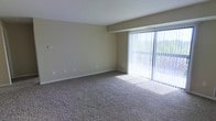 This single-floor residential space offers a clean, functional layout designed for comfortable everyday living. The main living area is a standout feature, showcasing wall-to-wall carpeting in a neutral tone that adds warmth underfoot and complements the soft beige walls throughout. A large sliding glass door dominates one wall of the living area, fitted with vertical blinds that allow for flexible light control. This expansive glazing floods the room with natural light and frames views of verdant, tree-lined surroundings, creating a pleasant connection between the interior and the outdoors. The open feel of the living space is enhanced by clean white ceilings and simple, unfussy finishes that lend the room a versatile, move-in-ready appeal. The neutral color palette of beige walls and tan carpeting creates a cohesive, calming ambiance throughout. Subtle architectural details such as clean baseboard trim and well-placed electrical outlets reflect a thoughtfully practical design. The overall atmosphere is bright, airy, and welcoming, making it well-suited as a comfortable personal retreat or an easily personalized living environment.