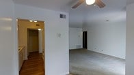 This single-floor residential space presents a straightforward and functional layout designed for comfortable everyday living. The main living area is generously sized, featuring wall-to-wall carpeting that adds warmth underfoot and a ceiling fan with a built-in light fixture that provides both air circulation and ambient illumination. Vaulted or angled ceilings give the space a sense of added volume and openness. Neutral white walls throughout create a bright, clean backdrop that allows for versatile interior styling. HVAC vents are visible in both the ceiling and near the floor, ensuring year-round climate control. Adjacent to the main living area, a corridor leads to a bedroom or secondary space, which is highlighted by a walk-in closet equipped with a hanging rod and overhead lighting — a practical storage feature. The flooring transitions from carpet in the living areas to hardwood or wood-look flooring in the connecting hallway, adding visual contrast and textural variety. The overall ambiance is minimal and move-in ready, with a neutral palette and practical finishes that suit a wide range of furnishing styles. This space offers a solid foundation for personalization and comfortable single-level living.