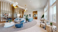 This expansive single-floor amenity space delivers a resort-inspired experience through thoughtful architecture and high-end interior design. The centerpiece of the space is a dramatic lounge area anchored by a circular area rug and terrazzo flooring, where a striking installation of angled wood slat columns rises toward the ceiling, creating a bold sculptural focal point. Oversized floral-shaped pendant lights in white and gold add warmth and elegance overhead. Seating areas are appointed with plush blue velvet chairs, soft neutral sofas, and accent pillows, evoking a relaxed yet sophisticated atmosphere enhanced by lush indoor plants and curated wall art.

Adjacent to the main lounge, a game room features multiple billiard tables, a high-top dining bar, and large windows that flood the area with natural light, offering views of the outdoors. Flowing white drapery and gold pendant lighting maintain a cohesive, upscale aesthetic throughout.

A dedicated co-working area offers a vibrant contrast with bold mustard-yellow velvet lounge chairs arranged beneath sculptural cluster chandeliers. Glass-partitioned private office rooms lined in black steel frames provide individual workspaces while preserving an open, airy feel.

A sleek wet bar rounds out the space, featuring a curved white countertop accented with blue tile, gold hardware, and pendant lighting, with tall windows framing outdoor greenery.