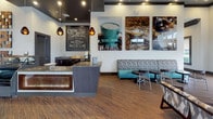 Located at 3115 Grand Ave in Pinellas Park, Florida, this single-floor commercial space serves as a stylish leasing office and resident lounge. The interior greets visitors with floor-to-ceiling glass entry doors and expansive windows that flood the space with natural light and frame views of the palm-lined exterior. Rich wood-look flooring runs throughout, complementing a contemporary color palette of charcoal, teal, and warm neutrals.

A prominent coffee bar anchors one side of the space, featuring a granite-topped counter with decorative mosaic tile paneling, pendant lighting, and a chalkboard menu board displaying espresso and coffee offerings. Large-format photography prints of coffee beans and beverages adorn the adjacent wall, reinforcing the café aesthetic. Comfortable banquette seating with patterned upholstery and teal accent chairs provide relaxed lounge areas for visitors.

Toward the rear, a high-top counter with bar stools runs along the window wall, offering a bright workspace with outdoor views. A dedicated workstation area with computer terminals serves leasing and administrative functions. Tray ceiling details with recessed lighting and dark accent trim add architectural depth, while roller shades allow for adjustable light control throughout the day. The overall ambiance is welcoming, modern, and professionally appointed.