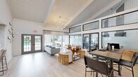 Located at 7910 Crossroads Dr in North Charleston, SC, this single-floor community space delivers a sleek, contemporary design with an airy, welcoming ambiance throughout. The main lounge area is an architectural highlight, boasting soaring vaulted ceilings clad in white shiplap, expansive clerestory windows that flood the interior with abundant natural light, and light-toned hardwood flooring that ties the space together. A striking sputnik chandelier serves as a focal point overhead, while floor-to-ceiling black-framed glass doors and windows connect the lounge to a dedicated fitness area, maintaining an open, cohesive feel. Double glass doors lead directly to an outdoor pool area, further enhancing the sense of openness and bringing the outdoors in. The fully appointed kitchen features quartz countertops with marble-patterned backsplash tile, cream cabinetry with matte black hardware, open shelving, and a window above the sink that frames lush greenery outside. Recessed lighting and a full-size refrigerator round out the kitchen's functionality. A casual dining area within the kitchen accommodates seating for small groups. Throughout, the palette of whites, soft neutrals, and natural wood tones creates a cohesive, sophisticated atmosphere ideal for gathering and relaxation.