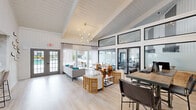 Located at 7910 Crossroads Dr in North Charleston, SC, this single-floor community space delivers a sleek, contemporary design with an airy, welcoming ambiance throughout. The main lounge area is an architectural highlight, boasting soaring vaulted ceilings clad in white shiplap, expansive clerestory windows that flood the interior with abundant natural light, and light-toned hardwood flooring that ties the space together. A striking sputnik chandelier serves as a focal point overhead, while floor-to-ceiling black-framed glass doors and windows connect the lounge to a dedicated fitness area, maintaining an open, cohesive feel. Double glass doors lead directly to an outdoor pool area, further enhancing the sense of openness and bringing the outdoors in. The fully appointed kitchen features quartz countertops with marble-patterned backsplash tile, cream cabinetry with matte black hardware, open shelving, and a window above the sink that frames lush greenery outside. Recessed lighting and a full-size refrigerator round out the kitchen's functionality. A casual dining area within the kitchen accommodates seating for small groups. Throughout, the palette of whites, soft neutrals, and natural wood tones creates a cohesive, sophisticated atmosphere ideal for gathering and relaxation.