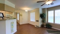 This single-floor home offers a practical and welcoming layout designed for comfortable everyday living. The main living area serves as the heart of the space, featuring warm wood-look hardwood flooring that extends throughout, creating a cohesive and inviting atmosphere. A ceiling fan overhead provides both airflow and a casual finishing touch to the room.

Natural light filters in through blinded windows, brightening the neutral-toned walls and giving the space an airy, open feel. The front entryway opens directly into the living area, providing a smooth and accessible flow from the moment you step inside.

Adjacent to the living space, the kitchen is partially visible and features a light-toned countertop, adding a clean contrast to the warm flooring. The open sightlines between the kitchen and living area support an interconnected layout ideal for both daily routines and entertaining.

The overall design of the home is straightforward and functional, with a neutral color palette of soft whites and warm beiges that lends itself well to a variety of interior styles. The single-floor layout ensures ease of movement throughout, making this a practical and comfortable living environment.