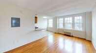 This single-floor residence combines classic architectural character with thoughtful modern updates throughout. The entryway greets visitors with warm hardwood flooring and clean white millwork, including crown molding and paneled doors with brushed nickel hardware, setting a refined tone for the rest of the space.

The kitchen is a standout feature, showcasing rich honey-toned wood cabinetry extending to the ceiling for ample storage, light speckled granite countertops, and a breakfast bar peninsula that opens to the living area. Black appliances, including a refrigerator and dishwasher, complement the cabinetry, while recessed lighting brightens the space. The kitchen flows seamlessly into the open living area, which is bathed in natural light from multiple large double-hung windows and features gleaming hardwood floors and a cast-iron radiator.

The bedrooms continue the hardwood flooring theme and are enhanced by crown molding, natural light from generously sized windows with outdoor views, and ceiling-mounted light fixtures. Exposed beam detailing adds architectural interest in select areas.

The bathroom is finished in a crisp, classic style with white subway tile surrounding the soaking tub and shower, a pedestal sink, penny tile flooring, and a window providing natural ventilation — a timeless and clean aesthetic throughout.