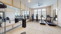 Located at 701 S Buncombe Rd in Greer, South Carolina, this fitness center is thoughtfully designed to support a comprehensive workout experience. The space is finished with large-format tile flooring throughout, neutral walls, and warm wood-toned trim accents that frame the room's large windows, flooding the interior with natural light and offering views of the surrounding community.

The cardio zone is well-appointed with multiple treadmills and elliptical machines positioned along the windowed wall, making workouts feel open and energizing. A ceiling fan overhead contributes to airflow and comfort during exercise. An expansive wall-mounted mirror runs the length of one side of the room, visually enlarging the space while allowing users to monitor their form. Flanking the mirror, a full dumbbell rack offers a range of free weights for strength training.

A dedicated cable machine with an adjustable bench, branded by BodyCraft, anchors the strength training area, providing versatile resistance options. A separate, quieter nook within the space features carpet flooring and a wall-mounted foldable mat, ideal for stretching, yoga, or recovery work. The fitness center is accessed through a glass-panel door and includes recessed lighting, creating a clean, modern atmosphere suitable for all fitness levels.