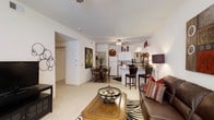 This single-floor residence is designed with an open-concept layout that seamlessly connects the main living areas. The living room offers a comfortable, well-proportioned space anchored by plush carpeting, a wood coffee table, and a ceiling fan overhead. Decorative accents, including wall art and a zebra-print area rug, add character to the space.

Adjacent to the living area, a defined dining space accommodates a round table with chairs, positioned beneath a second ceiling fan. The kitchen is bright and functional, featuring white cabinetry, a white refrigerator, and a breakfast bar with seating for additional casual dining. The bar-height counter provides a natural transition between the kitchen and the rest of the living space.

The layout flows efficiently, with a hallway leading to additional private areas of the home. White walls throughout contribute to a clean, airy aesthetic that maximizes the sense of space.

A private patio, framed by a classic white vinyl railing, extends the living space outdoors and overlooks a well-maintained landscape with mature pine trees, green lawns, and views of the broader community. The outdoor setting provides a peaceful retreat with an open, natural backdrop.