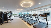 This single-level fitness center is designed with a clean, contemporary aesthetic that prioritizes both functionality and an open, inviting atmosphere. The space is organized into distinct workout zones, seamlessly connected by light wood and rubberized flooring that transitions appropriately between areas.

The cardio zone features a row of treadmills and elliptical trainers equipped with individual digital screens, positioned directly in front of expansive floor-to-ceiling glass walls. These large windows and sliding glass doors flood the space with natural light and provide unobstructed views of an outdoor terrace lined with tropical greenery, blue umbrellas, and lounge seating.

The strength training area is anchored by a power rack, cable machine, adjustable bench, and a medicine ball rack, complemented by a slatted wood accent wall and wall-mounted television. Overhead, distinctive circular LED light fixtures add a modern design element throughout.

A rowing machine and various additional cardio equipment, including a spin bike and stair climber, round out the offering. The facility also includes a dedicated locker room area featuring curved walls finished in blue mosaic tile, tiled flooring, and private changing rooms, providing a polished and complete amenity experience.