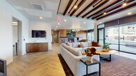 This thoughtfully designed single-floor amenity space blends contemporary style with functional comfort across several distinct areas. The centerpiece is a spacious communal lounge featuring striking herringbone hardwood floors, exposed dark wood beam ceilings, and a large-scale sectional sofa anchored by a dramatic floor-to-ceiling brick fireplace. Industrial-style steel-framed windows, a wall-mounted television, and warm ambient lighting contribute to an inviting, modern-industrial aesthetic throughout. A cozy seating nook adjacent to the fireplace offers wingback chairs, organic-shaped wood coffee tables, and framed artwork, creating an intimate retreat within the larger lounge. The fitness center is generously proportioned, offering a wide range of equipment including treadmills, cable machines, ellipticals, rowing machines, free weights, and a dedicated weight bench. A tray ceiling with recessed lighting and a ceiling fan keeps the space bright and comfortable, with large windows providing natural light and outdoor views. A separate yoga and stretching studio features soft carpeted flooring, dual ceiling fans, skylights, and ample open floor space with mats and accessories readily available. Completing the facility is a clean, well-organized mailroom with a tiled floor and a full bank of individual mailboxes, accessible via a dedicated entrance.