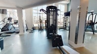 This fitness center is a thoughtfully designed, single-floor facility that offers a comprehensive workout environment for users of all training styles. The space is characterized by soaring ceilings and expansive floor-to-ceiling windows that flood the interior with natural light while providing views of an outdoor pool courtyard and the surrounding community.

The flooring throughout is polished concrete, providing a durable and clean aesthetic that complements the modern, functional design. Large mirrors are strategically placed to enhance the sense of space and support proper form during training.

The strength training area is well-appointed, featuring a multi-functional cable machine system, a Smith machine, weight benches, and a dedicated dumbbell rack stocked with a full range of free weights. A functional training zone includes a TKO punch bag, TRX suspension trainers, battle ropes, a pull-up station, and wall-mounted accessory storage, catering to high-intensity and functional fitness routines. A motivational "Train Hard" sign anchors this area visually.

The cardio section lines the windowed wall and includes treadmills, a stair climber, and additional cardio machines, all positioned to take advantage of the natural light and outdoor views. A wall-mounted television is also present for entertainment during workouts.

A separate corridor area houses an additional dual cable functional trainer, offering further versatility. Recessed lighting throughout ensures the space remains well-lit at all times.