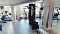 This fitness center is a thoughtfully designed, single-floor facility that offers a comprehensive workout environment for users of all training styles. The space is characterized by soaring ceilings and expansive floor-to-ceiling windows that flood the interior with natural light while providing views of an outdoor pool courtyard and the surrounding community.

The flooring throughout is polished concrete, providing a durable and clean aesthetic that complements the modern, functional design. Large mirrors are strategically placed to enhance the sense of space and support proper form during training.

The strength training area is well-appointed, featuring a multi-functional cable machine system, a Smith machine, weight benches, and a dedicated dumbbell rack stocked with a full range of free weights. A functional training zone includes a TKO punch bag, TRX suspension trainers, battle ropes, a pull-up station, and wall-mounted accessory storage, catering to high-intensity and functional fitness routines. A motivational "Train Hard" sign anchors this area visually.

The cardio section lines the windowed wall and includes treadmills, a stair climber, and additional cardio machines, all positioned to take advantage of the natural light and outdoor views. A wall-mounted television is also present for entertainment during workouts.

A separate corridor area houses an additional dual cable functional trainer, offering further versatility. Recessed lighting throughout ensures the space remains well-lit at all times.