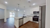 This single-story unit in Athens, Texas, offers a thoughtfully designed layout with modern finishes throughout. The open-concept living and kitchen area serves as the heart of the space, anchored by a substantial kitchen island with granite countertops, an integrated sink with a matte black faucet, and pendant lighting overhead. White shaker-style cabinetry lines the walls, complemented by a full suite of stainless steel appliances, including a refrigerator, electric range, over-the-range microwave, and dishwasher. Rich, dark-toned hardwood-style flooring runs continuously throughout the unit, providing a warm contrast to the bright white walls and trim. The living area features sliding glass doors that open onto a private balcony, allowing natural light to fill the space and providing outdoor views. The bedroom is a well-proportioned, serene retreat with a ceiling fan, a window with blinds, and direct access to the bathroom. The bathroom features a granite-topped vanity with white cabinetry, a large mirror, and a full soaking tub with classic white subway tile surround. Additional storage is available through a dedicated closet space, and a utility area accommodates laundry connections.