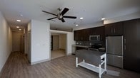 This single-floor residence offers a clean, modern aesthetic with an open-concept layout that seamlessly connects the kitchen and living areas. The kitchen is well-appointed with rich, dark espresso cabinetry that provides ample storage, complemented by a tile backsplash and stainless steel appliances including a refrigerator, electric range, and built-in microwave. A mobile kitchen island on casters adds versatile additional prep and storage space. Recessed lighting throughout the kitchen ensures bright, even illumination, while a ceiling fan in the living area promotes comfortable airflow. The consistent hardwood-style flooring flows throughout the open living space, contributing to a cohesive and contemporary feel. A hallway visible from the main living area leads to additional rooms, including what appears to be a walk-in closet or pantry space. The interior features crisp white walls that contrast with the dark cabinetry, creating a bold yet inviting atmosphere. The thoughtful combination of functional design elements and modern finishes makes this a highly practical and stylish living space.