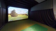This single-level indoor golf simulator space is thoughtfully designed to deliver an immersive, year-round golfing experience. At the heart of the room is a large, high-definition projection screen that displays detailed, lifelike golf course visuals, complete with on-screen course mapping and scoring interfaces. Dark acoustic curtains line the walls surrounding the simulator bay, minimizing distractions and optimizing the visual environment for play.

The flooring throughout the hitting area is covered in artificial turf in varying shades of green, replicating a realistic course feel underfoot. A dedicated hitting mat with tee markers is positioned directly in front of the screen, providing an authentic stance and swing zone. Overhead, exposed dark beams and recessed lighting contribute to the room's focused, purposeful ambiance.

Adjacent to the simulator bay, a comfortable lounge area features rich brown leather armchairs and side tables, offering a relaxed space for players to rest between rounds or observe others. A wall-mounted control panel and club storage rack are conveniently positioned nearby for easy access during sessions.

A glass-paneled entry door connects the simulator room to a warmly lit reception or common area beyond, suggesting this space is part of a larger facility. Overall, the room balances technical sophistication with comfortable, club-like aesthetics.