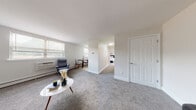 This single-floor residence offers a thoughtfully designed layout that balances functionality with a clean, neutral aesthetic throughout. The living area is carpeted in soft gray and receives natural light through multiple windows fitted with blinds, creating a bright and inviting atmosphere. A wall-mounted air conditioning unit and baseboard heating provide year-round climate control. The space flows from the living area into a defined dining zone and continues toward the bedroom through a hallway.

The kitchen is well-appointed with white shaker-style cabinetry extending to the ceiling, granite-look countertops, a white subway tile backsplash, and a full suite of stainless steel appliances including a refrigerator, electric range, dishwasher, and microwave. Light wood-tone vinyl flooring adds warmth to this functional galley-style space, which also accommodates a small dining table near a window.

The bedroom is comfortably sized with plush carpet, a closet with standard rod storage, and access to an adjacent bathroom. The bathroom features light wood-tone tile flooring, a white vanity with modern gold-toned hardware, and grab bars, indicating accessibility-conscious design. Overall, the unit presents a cohesive, move-in-ready environment with modern finishes throughout.
