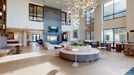 Located at 2168 Fox Valley Center in Aurora, IL, this contemporary community amenity space makes an immediate impression with its grand, double-height atrium-style lobby. The centerpiece of the main hall is a sweeping circular sectional seating arrangement surrounding a live planter, all beneath a breathtaking cascading floral chandelier that draws the eye upward toward the expansive clerestory windows that flood the space with natural light.

A floor-to-ceiling slate-toned fireplace anchors one wall, paired with a mounted flat-screen display and flanked by clean-lined upholstered seating. Light wood-toned flooring runs throughout, unifying the open-plan layout and contributing to the airy, contemporary aesthetic.

Transitioning deeper into the space, a second lounge area features generous deep-blue sectional sofas, a sleek mixed-material coffee table, and a circular pendant light fixture, creating a more intimate yet equally stylish setting. Glass partitions and steel-framed windows maintain visual connectivity between zones.

A separate sunlit room with exposed light brick walls, soaring ceilings, globe pendant lighting, and French doors opening to an outdoor terrace provides a bright, casual gathering area with woven lounge seating. Throughout, the design balances modern sophistication with comfort, making this a highly functional and visually compelling communal destination.