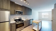 This single-floor unit showcases a contemporary design with clean lines and a neutral color palette throughout. The kitchen serves as a standout feature, outfitted with dark wood-grain cabinetry extending to the ceiling for generous storage, stainless steel appliances including a gas range, over-the-range microwave, refrigerator, and dishwasher, along with light quartz countertops and a subtle subway tile backsplash. A track lighting fixture illuminates the workspace, and light-toned hardwood-style flooring ties the kitchen into the adjacent living area seamlessly.

The living space is bright and open, benefiting from natural light and connecting directly to a carpeted bedroom. The bedroom features a sliding glass door fitted with vertical blinds, providing access to an outdoor space and views of the surrounding area, while a separate window adds additional daylight.

The full bathroom is finished with large-format tile flooring, a dark wood-grain vanity topped with a white quartz surface, a wide mirror with overhead lighting, and a tub-shower combination with vertically tiled surround walls. The overall ambiance of the unit is modern, clean, and move-in ready.