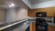 This two-story space is designed with practicality and comfort in mind. The main level features carpeted flooring throughout, lending a soft, warm feel to the living areas. A staircase with a white-painted balustrade and a wooden handrail connects both floors, positioned alongside a hallway that leads to additional rooms. Natural light filters into the main living area through draped sliding glass doors, offering access to an outdoor space.

The kitchen is well-appointed with warm honey-toned wood cabinetry providing ample upper and lower storage. Dark speckled countertops extend generously across the workspace, accommodating a double-basin stainless steel sink. Black appliances, including an electric range and an over-the-range microwave, complete the functional cooking space. Recessed lighting above the kitchen area ensures the workspace is well illuminated. The neutral gray tones used throughout the walls create a cohesive, understated aesthetic across both levels, providing a versatile backdrop that complements a variety of interior styles.