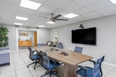 Located at 4156 NW 21st Ave in Oakland Park, Florida, this professional conference room offers a functional and well-designed single-floor layout ideal for meetings and collaborative gatherings. The space is anchored by a substantial light wood conference table surrounded by ergonomic blue mesh chairs, accommodating a sizable group comfortably. A wall-mounted flat-screen display with an integrated camera enhances the room's video conferencing capabilities, making it well-suited for modern business needs.

The room features a drop-panel ceiling with recessed fluorescent lighting panels that provide bright, even illumination throughout, complemented by a ceiling fan that promotes air circulation. Light-toned large-format ceramic tile flooring runs throughout, lending the space a clean, professional aesthetic. Natural light filters in through a window fitted with horizontal blinds, adding warmth to the interior.

A dedicated lounge area along one wall includes a plush sofa and a pair of patterned accent chairs with a side table, offering a relaxed setting for informal discussions. Decorative framed artwork adorns multiple walls, adding color and character to the otherwise neutral palette. Additional storage and a credenza are visible toward the back of the room, contributing to the space's overall functionality and polished presentation.