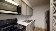 This single-floor residence offers a well-organized layout with distinct living, kitchen, and bedroom areas connected by wood-look flooring throughout the main spaces. The kitchen is efficiently designed with gray shaker-style cabinetry, light speckled countertops, a stainless steel double-basin sink, and a full suite of Frigidaire appliances including an electric range, over-the-range microwave, and refrigerator. A textured ceiling adds character to the kitchen area. The open living room is a standout space, featuring rich, dark wood-look flooring and a stone-surround fireplace that serves as a natural focal point. A sliding glass door fills the room with natural light and provides access to an outdoor area, offering views of mature trees. A hallway connects the living areas to the private quarters, where carpeted bedrooms provide a softer underfoot feel. The bedrooms are bright, with windows fitted with blinds that allow for controlled natural light. The bathroom features wood-look flooring consistent with the rest of the home, a vanity with light countertops, and a clean, straightforward layout. Overall, the space presents a practical and cohesive design with a neutral color palette throughout.