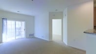 This single-floor space presents a clean, minimalist aesthetic defined by bright white walls and soft neutral carpeting throughout. The main living area is generously proportioned, offering an open feel enhanced by natural light entering through a glass-paneled door, likely leading to an outdoor area. The room's height and unobstructed layout create an airy, welcoming atmosphere.

A wide doorway connects the main living space to an adjacent room, suggesting a flexible floor plan well-suited for varied uses. Floor vents are positioned throughout, indicating a central HVAC system for year-round climate control. The absence of furniture highlights the clean lines of the architecture and the seamless transitions between spaces.

The overall condition of the space appears well-maintained, with smooth ceilings, clean baseboards, and evenly finished walls. The consistent flooring and neutral color palette provide a versatile backdrop adaptable to a range of interior design styles. The layout emphasizes functionality and flow, making efficient use of the available footprint while maintaining a bright, open ambiance throughout.
