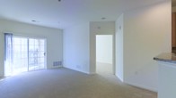 This single-floor space presents a clean, minimalist aesthetic defined by bright white walls and soft neutral carpeting throughout. The main living area is generously proportioned, offering an open feel enhanced by natural light entering through a glass-paneled door, likely leading to an outdoor area. The room's height and unobstructed layout create an airy, welcoming atmosphere.

A wide doorway connects the main living space to an adjacent room, suggesting a flexible floor plan well-suited for varied uses. Floor vents are positioned throughout, indicating a central HVAC system for year-round climate control. The absence of furniture highlights the clean lines of the architecture and the seamless transitions between spaces.

The overall condition of the space appears well-maintained, with smooth ceilings, clean baseboards, and evenly finished walls. The consistent flooring and neutral color palette provide a versatile backdrop adaptable to a range of interior design styles. The layout emphasizes functionality and flow, making efficient use of the available footprint while maintaining a bright, open ambiance throughout.