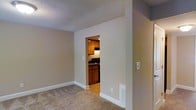Located at 1933 North Hills Dr in Raleigh, NC, this single-story home presents a straightforward and functional layout designed for comfortable everyday living. The living area is finished with wall-to-wall carpeting in a warm neutral tone, complemented by crisp white baseboards and smooth beige walls that create a clean, understated aesthetic throughout. A textured popcorn ceiling adds a classic touch overhead, while a floor vent ensures efficient climate control.

An open doorway connects the living space directly to the kitchen, offering a sense of flow between the two areas. The kitchen features warm-toned wood cabinetry providing ample storage, dark countertops, and hardwood-style flooring that contrasts with the carpeted living space. An under-cabinet range hood is visible above the cooktop, and recessed lighting illuminates the kitchen's compact yet functional footprint.

The neutral color palette carried throughout the home creates a cohesive and inviting atmosphere, making the space easy to personalize. The single-floor layout offers accessibility and convenience, with everything situated on one level for seamless movement through the home.