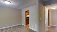 Located at 1933 North Hills Dr in Raleigh, NC, this single-story home presents a straightforward and functional layout designed for comfortable everyday living. The living area is finished with wall-to-wall carpeting in a warm neutral tone, complemented by crisp white baseboards and smooth beige walls that create a clean, understated aesthetic throughout. A textured popcorn ceiling adds a classic touch overhead, while a floor vent ensures efficient climate control.

An open doorway connects the living space directly to the kitchen, offering a sense of flow between the two areas. The kitchen features warm-toned wood cabinetry providing ample storage, dark countertops, and hardwood-style flooring that contrasts with the carpeted living space. An under-cabinet range hood is visible above the cooktop, and recessed lighting illuminates the kitchen's compact yet functional footprint.

The neutral color palette carried throughout the home creates a cohesive and inviting atmosphere, making the space easy to personalize. The single-floor layout offers accessibility and convenience, with everything situated on one level for seamless movement through the home.