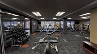 This fitness center is a comprehensively outfitted exercise facility designed to support a wide range of workout routines. The space is organized into distinct functional zones, providing users with ample room to move between equipment comfortably. The flooring features a patterned commercial carpet with orange and teal accent tiles that define workout areas throughout the space.

Along the windowed wall, a row of treadmills and elliptical machines faces large picture windows that allow natural light to flow in and provide views of the surrounding exterior — an energizing backdrop for cardio sessions. Mounted flat-screen televisions are positioned nearby for added entertainment. Additional cardio options, including upright bikes and stair climbers, are arranged in an adjacent area alongside the window line.

The free weights section is equipped with a full rack of dumbbells and is positioned near a large floor-to-ceiling mirror, enhancing the sense of space and allowing for form monitoring during workouts. Adjustable benches and cable resistance machines are distributed throughout, supporting strength and functional training. A multi-station cable machine anchors the center of the room.

A water fountain and waste receptacle are conveniently accessible near the entry. The drop ceiling with recessed panel lighting ensures consistent, even illumination throughout the entire facility.