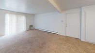 This single-floor unit offers a straightforward and functional layout across one bedroom and one bathroom. The living area is generously proportioned, featuring neutral-toned wall-to-wall carpeting and white walls that create a bright, open feel. A wall-mounted air conditioning unit and baseboard heating provide year-round climate control, while vertical blinds along a wide window allow for natural light management. The space opens directly into a separate kitchen, distinguished by tile flooring, warm laminate countertops, white cabinetry, and a full suite of white appliances including a gas range with overhead ventilation, a dishwasher, and a refrigerator. A stainless steel sink anchors the counter space. The bedroom continues the carpeted flooring found throughout the living areas and includes a double-door closet with built-in shelving and a hanging rod for ample storage. The full bathroom is finished with classic pink ceramic tile surrounding a shower-tub combination, a mosaic tile floor, a white vanity with countertop sink, and a ventilation fan. The overall aesthetic is clean and utilitarian, well-suited for straightforward, comfortable living.
