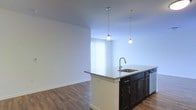 This single-floor space showcases a clean, modern design centered around an open-concept layout that seamlessly connects the kitchen and living areas. The kitchen features a prominent island with a light stone countertop, an undermount sink with a contemporary pull-down faucet, and dark espresso cabinetry with sleek hardware. Two pendant lights hang above the island, providing focused task lighting while contributing to the space's modern aesthetic. The cabinetry extends along the base of the island, offering ample storage. Adjacent to the kitchen, the living area is finished with warm wood-style flooring that runs throughout, creating a cohesive and inviting atmosphere. Crisp white walls and high ceilings amplify the sense of openness, while a large window dressed with vertical blinds allows generous natural light to fill the space. The overall ambiance is contemporary and well-lit, with a neutral color palette that provides a versatile backdrop for a variety of furnishing styles.