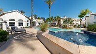 Located at 4630 S Lakeshore Dr in Tempe, Arizona, this resort-style outdoor amenity area showcases a generously sized, freeform swimming pool as its centerpiece. The crystal-clear, turquoise water is complemented by decorative blue tile accents along the pool's edge, adding a refined touch to the overall aesthetic. Surrounding the pool, expansive travertine-tiled decking provides ample space for lounge chairs and outdoor seating, ideal for relaxation under the abundant Arizona sunshine. Towering palm trees are thoughtfully distributed throughout the space, offering natural shade and reinforcing the tropical, resort-like atmosphere. The area is framed by Mediterranean-inspired architecture featuring stucco exteriors, arched windows, and terracotta tile rooflines, creating a cohesive and visually appealing backdrop. Raised planter beds with desert-adapted landscaping add texture and greenery throughout the deck area. Water features integrated into the pool design enhance the ambiance with gentle cascading elements. Shaded seating areas with patio umbrellas are positioned around the perimeter, offering comfortable retreats. The open-air setting benefits from unobstructed views of the clear blue Arizona sky, making this outdoor space a standout communal feature well-suited to the warm Tempe climate.