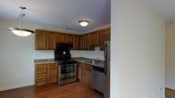 This single-floor residential space is designed with practicality and comfort in mind. The kitchen serves as a central highlight, outfitted with a full suite of stainless steel appliances including a gas range with oven, built-in dishwasher, and a full-size refrigerator. Rich, warm-toned wood cabinetry lines the walls, offering generous storage with upper and lower cabinet configurations that maximize the available space. The countertops feature a granite-style surface that complements the cabinetry, while a stainless steel sink with a single-handle faucet is conveniently positioned beneath additional cabinet storage. A black range hood is mounted above the cooktop, providing functional ventilation. The kitchen floor is finished in hardwood, adding a warm and classic aesthetic to the space. Flush-mount ceiling lighting provides even illumination throughout the kitchen. The layout follows an L-shaped configuration, allowing for an efficient workflow between the cooking, prep, and cleaning zones. The overall ambiance is cozy and functional, with traditional design elements that create a welcoming, lived-in feel suited for everyday use.