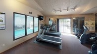 Located at 636 E Magnesium Rd in Spokane, Washington, this fitness center is a well-equipped, single-floor facility designed to support a comprehensive workout routine. The space is organized into distinct exercise zones, offering a variety of cardio and strength training options. A row of treadmills lines one side of the room, positioned beneath a wall-mounted television and accompanied by views of an adjacent indoor pool through large windows, creating a bright and motivating environment. A sliding glass door provides direct access to an outdoor patio area, allowing natural light to fill the space. The cardio section is further complemented by stationary bikes and an elliptical machine, catering to a range of fitness preferences. A dedicated strength training area houses a multi-station cable machine, a weight bench, and additional resistance equipment, all positioned in front of full-length wall mirrors that enhance the sense of space and allow users to monitor their form. The flooring throughout is a durable, dark carpet suited for gym use. Track lighting and recessed ceiling fixtures provide consistent illumination across both zones. Vaulted ceilings in portions of the room contribute to an open, airy feel. Decorative potted plants add a touch of warmth to the otherwise functional setting.