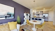 Located at 451 2nd Ave S in the heart of downtown Saint Petersburg, Florida, this contemporary residence offers a thoughtfully designed single-floor layout with a striking blend of modern aesthetics and functional living spaces. Light wood-style flooring flows throughout the open-concept living, dining, and kitchen areas, creating a cohesive and inviting atmosphere. Bold deep-purple accent walls add a dramatic yet sophisticated touch to both the living room and bedrooms.

The open kitchen is a standout feature, equipped with stainless steel appliances, sleek gray wood-grain cabinetry, dark stone countertops, and a peninsula with bar seating — ideal for casual dining or entertaining. Pendant lighting above the kitchen and dining area adds warmth and style.

The living area offers generous natural light through large windows with city views, complemented by contemporary furnishings and curated artwork. The primary bedroom features sliding glass doors opening to a private balcony overlooking the Saint Petersburg skyline, while a second bedroom showcases a dramatic dark accent wall with hanging pendant lights and a stacked mirror arrangement. The bathroom connects to an organized walk-in closet and features wood-look tile flooring and modern gray vanity cabinetry.