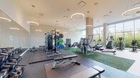 Located at 900 Pacific St in Stamford, Connecticut, this contemporary fitness center is a well-appointed facility designed to support a comprehensive workout experience. The space is organized into distinct functional zones, all unified by soaring ceilings, a neutral warm color palette, and oversized floor-to-ceiling windows that flood the interior with natural light and offer views of the surrounding greenery and cityscape.

The main workout floor features a robust selection of Matrix strength machines, a multi-rack cable system loaded with bumper plates, adjustable benches, and multiple tiers of dumbbells neatly arranged on dedicated racks. Turf flooring sections define the functional training area, which houses additional resistance machines and rowing equipment. A row of Matrix treadmills and cardio machines lines the perimeter, complemented by upright and recumbent bikes. A built-in wooden cubby storage unit provides convenient towel and accessory storage.

A separate studio room offers a bright, open setting with cushioned flooring, full-length mirrored walls, a wall-mounted display screen, and speakers — ideal for group classes or personal training. An adjacent room provides functional training accessories including stability balls, kettlebells, resistance tools, and plyo boxes. A water fountain station is also available on the main floor. Circular pendant lighting throughout adds a modern, polished aesthetic to the entire facility.