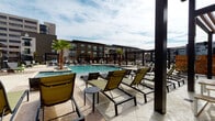 This thoughtfully designed outdoor amenity area offers a resort-inspired experience centered around a large, sparkling pool surrounded by a generous sun deck finished in textured stone pavers. Rows of sleek, olive-toned lounge chairs with dark metal frames are arranged throughout the deck, providing ample space for relaxation under open skies. Towering palm trees and manicured landscaping add a vibrant, tropical touch to the environment, while wrought iron fencing defines the perimeter with an elegant yet secure boundary.

Adjacent to the pool deck, a covered outdoor pavilion serves as a versatile gathering space. The structure features rich warm-toned wood slat ceilings and accent walls, complemented by bold dark steel posts and columns that frame open sightlines to the surrounding grounds. Ceiling fans ensure comfort throughout the warmer months. Built-in countertops with granite surfaces and grilling stations line one wall, making the space ideal for outdoor cooking and entertaining. Slatted wood picnic-style tables and benches provide communal seating for dining and socializing.

The overall design blends contemporary architectural materials — including brick, metal, and natural wood — with lush greenery and open-air connectivity, creating an inviting and functional outdoor retreat.