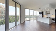 This contemporary high-rise residence showcases a sleek, modern design highlighted by dramatic floor-to-ceiling glass walls that flood the open-concept living and dining area with natural light while offering expansive views of the surrounding cityscape and lush treetops. Rich hardwood flooring flows throughout the main living spaces, complementing the clean white walls and recessed lighting overhead.

The chef's kitchen is a standout feature, appointed with striking quartz countertops with bold veining, a large waterfall island with an undermount sink and matte black faucet, dual-tone cabinetry in white and dark finishes, stainless steel appliances including a gas range, and a built-in wine refrigerator — all framed by floor-to-ceiling windows overlooking the outdoors.

The bedrooms are carpeted for comfort, each bathed in natural light through large windows with urban and greenery views. One bedroom includes a spacious walk-in closet with built-in shelving. The bathrooms are elegantly finished with marble-look tile flooring, dark-cabinet vanities with white quartz countertops, and glass-enclosed showers. One full bath also includes a soaking tub. A powder room with a pedestal sink completes the layout. The overall ambiance is bright, refined, and contemporary.