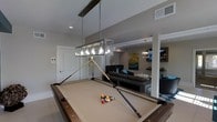 Located at 3101 Rise Dr in Morrisville, NC, this single-story community clubhouse is designed for social gatherings and leisure. The space showcases a modern, open-concept layout with a neutral color palette of warm grays and crisp whites that creates a clean, contemporary atmosphere throughout.

At the heart of the space is a full-size billiards table, elegantly lit by a statement rectangular pendant light with an industrial-chic glass and metal frame. The table serves as a natural focal point and anchor for the entertainment area.

Flowing seamlessly from the billiards zone is a comfortable lounge area, furnished with a plush sectional sofa and accented with vibrant teal throw pillows that add a pop of color. A wet bar or kitchenette area is visible in the background, providing convenient beverage service for gatherings. The lounge is further enhanced by a large-scale piece of wall art featuring blue and green tones, complementing the overall color scheme.

Additional seating and a mounted flat-screen television are visible deeper in the space, offering multiple zones for relaxation and entertainment. Warm pendant lighting and recessed ceiling lights provide layered illumination throughout. Tile flooring with a geometric-patterned area rug grounds the lounge area, adding texture and visual interest. Restroom facilities are also accessible directly from the main space.