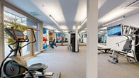 This modern fitness center offers a well-organized, single-floor exercise space designed to support a wide range of workout routines. The layout is open and spacious, with a neutral-toned carpet flooring and crisp white walls that create a clean, energizing atmosphere throughout.

The cardio zone is positioned along a wall of large floor-to-ceiling windows, allowing abundant natural light to fill the space and providing views of the surrounding outdoor area. This section includes treadmills, elliptical trainers, and cycling machines, complemented by a motivational "WORKOUT" wall graphic. Potted plants add a refreshing touch near the windows.

At the center of the space, a multi-use cable strength machine serves as an anchor for resistance training. Full-length mirrors line one wall, enhancing the sense of depth and supporting proper form during workouts. A wall-mounted flat-screen television is positioned for visibility from multiple areas of the room.

The functional training area houses a rack of exercise balls in varying sizes, additional stationary bikes, and open floor space suitable for stretching or mat work. Light wood shelving units provide storage for accessories and equipment. Recessed linear lighting fixtures run along the ceiling, ensuring the space is brightly and evenly illuminated. The overall design is modern, practical, and well-suited for a comprehensive fitness experience.