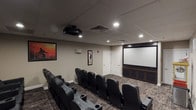 This private theater is designed to deliver an authentic cinematic experience within a well-appointed, dedicated screening room. The space is lined with rows of plush, high-back leather seats arranged in a tiered, stadium-style layout, each equipped with built-in cup holders for added comfort. Seating is organized to ensure unobstructed sightlines to the large projection screen mounted at the front of the room, which is supported by an overhead projector and a dark wood media console beneath the screen.

The room features a drop tile ceiling with recessed lighting and integrated surround sound speakers mounted along the walls, ensuring quality audio throughout. Decorative film reel accents and a curated selection of framed classic movie posters — including iconic titles — adorn the neutral-toned walls, reinforcing the cinematic atmosphere. Patterned carpet flooring adds warmth and acoustic dampening to the space.

A standout feature is the vintage-style popcorn cart stationed at the rear of the room, adding a charming, old-Hollywood touch. Two access doors are located near the screen wall for convenient entry and exit. Overall, the theater combines functional design with thoughtful décor to create an inviting and immersive movie-viewing environment.