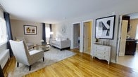 This single-floor space offers a bright and welcoming living area characterized by warm, honey-toned hardwood floors that extend throughout. The living room is thoughtfully arranged with a tufted gray sofa, patterned accent chairs, and a mirrored sunburst wall decoration as a focal point, lending the space a polished, contemporary aesthetic. A glass-top side table and area rug anchor the seating arrangement, while framed artwork on the walls adds visual interest. Natural light filters through windows dressed with dark gray curtains, complementing the soft, neutral wall palette. A white dresser with ornate detailing and decorative accessories is positioned near large framed art, adding a refined touch. Two interior doors lead to additional rooms, including what appears to be a bedroom visible in the background, suggesting a functional and connected layout. The space maintains a clean, cohesive design throughout, with white trim and moldings providing crisp contrast against the warm flooring and neutral walls. Overall, the space exudes a calm, livable ambiance enhanced by carefully chosen furnishings and decor.
