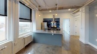 This single-floor urban loft blends industrial character with modern updates to create a distinctive and functional living space. Polished concrete floors run throughout, reflecting light and reinforcing the space's contemporary aesthetic. The open-plan living and kitchen area is anchored by a striking kitchen island finished in a muted blue-gray tone, topped with light countertops and equipped with a built-in sink. The kitchen features stainless steel appliances, including a refrigerator and electric range, complemented by a bold deep-blue subway tile backsplash, open floating shelves, and a stainless steel hood vent. Pendant Edison bulb fixtures hang from the exposed ceiling, which showcases raw beams, piping, and industrial track lighting — hallmarks of loft-style architecture. Large double-hung windows with black roller shades flood the space with natural light and offer elevated views of the surrounding cityscape, while cast-iron radiators beneath the windows nod to the building's historic roots. A dedicated bedroom area provides a quieter retreat, also benefiting from generous window coverage and city views. The bathroom is clean and minimal, featuring a glass-enclosed walk-in shower, standard fixtures, and a simple, utilitarian finish consistent with the overall design language of the space.