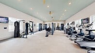 Located at 5251 W Nine Mile Rd in Pensacola, Florida, this single-floor fitness center offers a generously sized exercise space designed to accommodate a wide variety of workout routines. The room features soaring vaulted ceilings painted in a soft seafoam green, recessed lighting, and dual ceiling fans that contribute to a bright and comfortable atmosphere. Large windows along the perimeter flood the space with natural light while providing views of the surrounding exterior.

The facility is comprehensively outfitted with an extensive selection of equipment. Strength training is well-represented with cable machines, multi-station weight machines, squat racks, barbells, and a full tiered dumbbell rack. Cardiovascular options include treadmills, stair climbers, rowing machines (including a RowERG), and stationary bikes. A dedicated functional training area features stability balls, resistance bands, medicine balls, and a medicine ball storage rack.

The interior design blends clean white walls with warm wood-panel accent features and large framed mirrors that enhance the sense of space. Built-in cabinetry with white cabinet doors provides additional storage, while wall-mounted televisions are positioned throughout for entertainment during workouts. A water fountain station with green tile accents adds a practical finishing touch to this modern, well-appointed fitness facility.