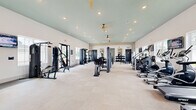 Located at 5251 W Nine Mile Rd in Pensacola, Florida, this single-floor fitness center offers a generously sized exercise space designed to accommodate a wide variety of workout routines. The room features soaring vaulted ceilings painted in a soft seafoam green, recessed lighting, and dual ceiling fans that contribute to a bright and comfortable atmosphere. Large windows along the perimeter flood the space with natural light while providing views of the surrounding exterior.

The facility is comprehensively outfitted with an extensive selection of equipment. Strength training is well-represented with cable machines, multi-station weight machines, squat racks, barbells, and a full tiered dumbbell rack. Cardiovascular options include treadmills, stair climbers, rowing machines (including a RowERG), and stationary bikes. A dedicated functional training area features stability balls, resistance bands, medicine balls, and a medicine ball storage rack.

The interior design blends clean white walls with warm wood-panel accent features and large framed mirrors that enhance the sense of space. Built-in cabinetry with white cabinet doors provides additional storage, while wall-mounted televisions are positioned throughout for entertainment during workouts. A water fountain station with green tile accents adds a practical finishing touch to this modern, well-appointed fitness facility.