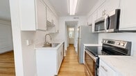 This 510-square-foot, one-bedroom, one-bathroom apartment at 3227 S Parker Rd in Denver offers a functional single-floor layout. The open living area features light wood-style flooring and white walls, creating a bright, clean aesthetic. The galley-style kitchen is equipped with white shaker cabinets, light stone countertops, stainless steel appliances including a refrigerator, electric range, and microwave, plus a sink and ample cabinet storage. The 130-square-foot bedroom features plush carpet, a natural light-admitting window, and a closet with built-in shelving. The bathroom includes a full tub/shower combo, vanity with white cabinetry, and wood-style flooring. A dedicated laundry area and hallway complete the layout.