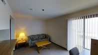 This single-floor space offers a straightforward and functional layout centered around a comfortable living area. The living room is finished with wall-to-wall carpeting that adds warmth underfoot, and features neutral-toned walls that create a clean, understated backdrop. A large sliding glass door fitted with vertical blinds serves as the room's primary source of natural light, allowing brightness to filter in while maintaining privacy. The seating arrangement includes a patterned sofa with a classic design, accompanied by a wooden folding coffee table that provides practical surface space. A side table with a lamp offers additional ambient lighting, contributing to a relaxed atmosphere. The smooth, flat ceiling is equipped with a smoke detector and security camera, reflecting attention to safety. The overall layout is open and uncluttered, making the space easy to navigate and adaptable to various needs. The neutral palette and simple finishes give the space a calm, no-frills character well-suited for everyday living.