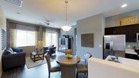 This single-floor residence showcases an open-concept design that integrates the dining and living areas into one cohesive, flowing space. The dining area is anchored by a round wooden table surrounded by leather-upholstered chairs, illuminated by an elegant pendant light with a brushed nickel finish. Adjacent to the dining space, the living area features a ceiling fan, complementing the natural light that filters in through sliding glass doors dressed with patterned curtains. The soft blue-grey wall tones create a calm, neutral backdrop throughout, enhanced by carefully chosen décor including a textured wall panel and decorative cabinetry. Dark hardwood-style flooring runs continuously through the space, grounding the contemporary aesthetic. The kitchen, visible from the dining area, features dark cabinetry and stainless steel appliances, partially framed by a white countertop overhang that defines the boundary between spaces. The overall ambiance is polished and modern, with layered lighting, curated furnishings, and a cohesive color palette that gives the space a welcoming yet sophisticated character.
